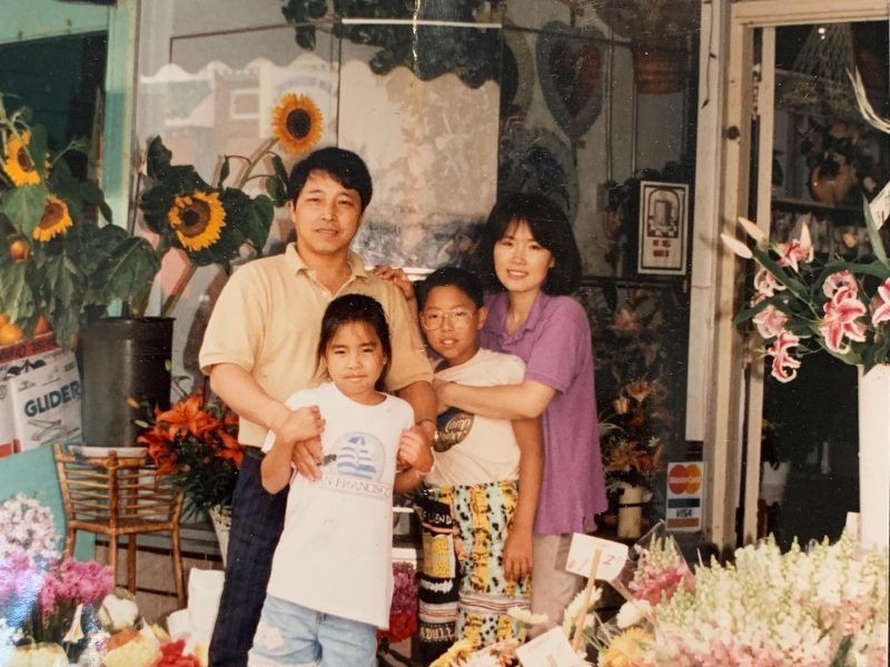A family of four stands together in front of a flower shop, with sunflowers and various floral arrangements around them.