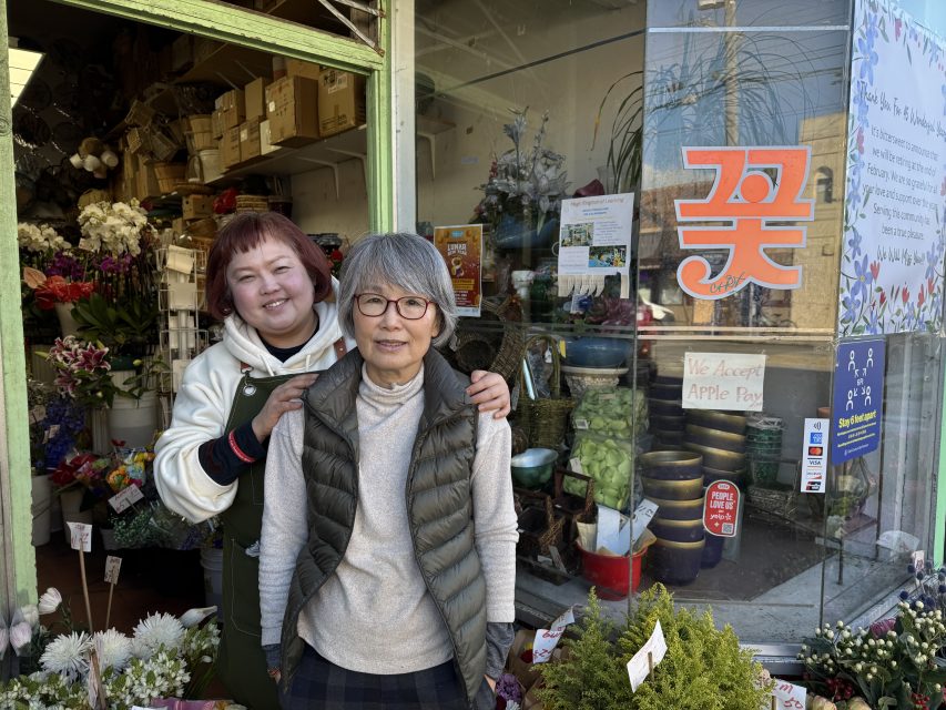 Two women stand in front of a flower shop filled with plants and flowers. One woman has her arms on the other's shoulders. A sign reading "We Accept Apple Pay" is visible on the glass door.