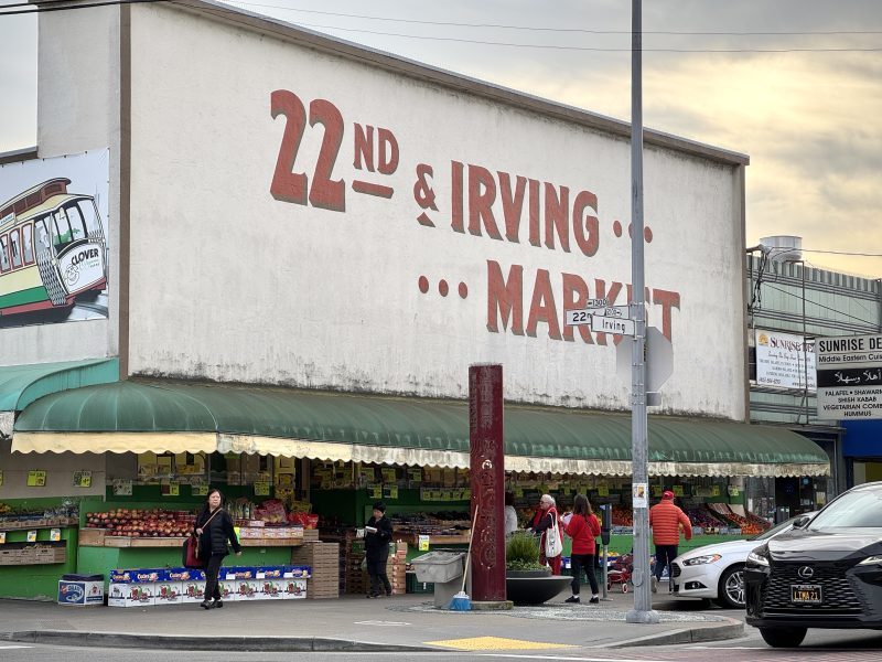 A market with a green awning stands at the corner of 22nd and Irving. Shoppers are passing by the storefront. The building has large painted letters reading "22nd & Irving Market.