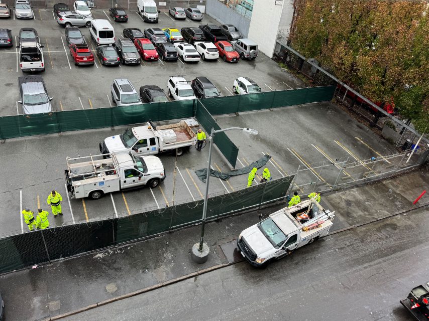 Workers on 6th Street, clad in high-visibility gear, install fencing around a parking lot with utility trucks parked nearby.