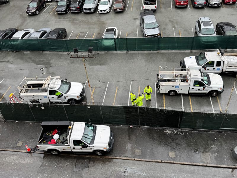 Aerial view of utility trucks lined up beside a fenced area on 6th Street, with several workers in hi-vis clothing standing and talking on a bustling construction site.