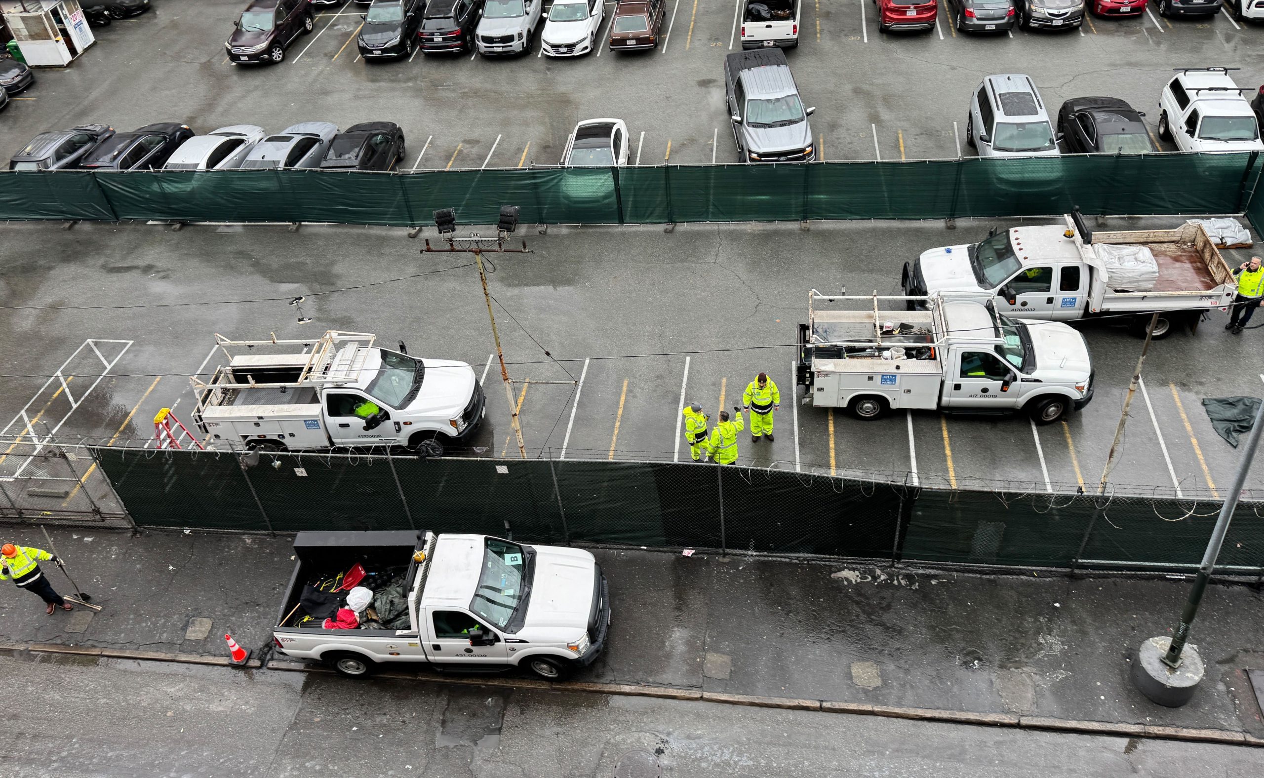 Aerial view of utility trucks lined up beside a fenced area on 6th Street, with several workers in hi-vis clothing standing and talking on a bustling construction site.