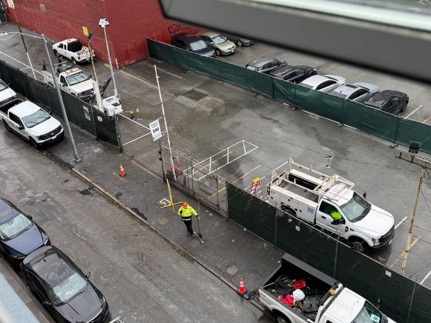 Aerial view of a construction site along 6th Street, bustling with workers, a white utility truck, and safety barriers. Vehicles line the adjacent street while a lot is visible behind a green fence.