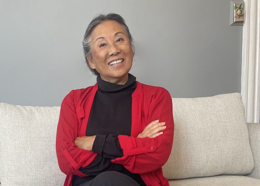A person with gray hair smiles while sitting on a light-colored sofa, wearing a black shirt and red cardigan, arms crossed.