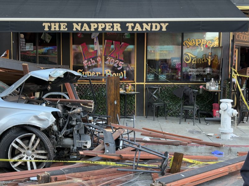 A damaged car and shattered wooden barriers rest outside The Napper Tandy bar, adjacent to a fire hydrant on the sidewalk.