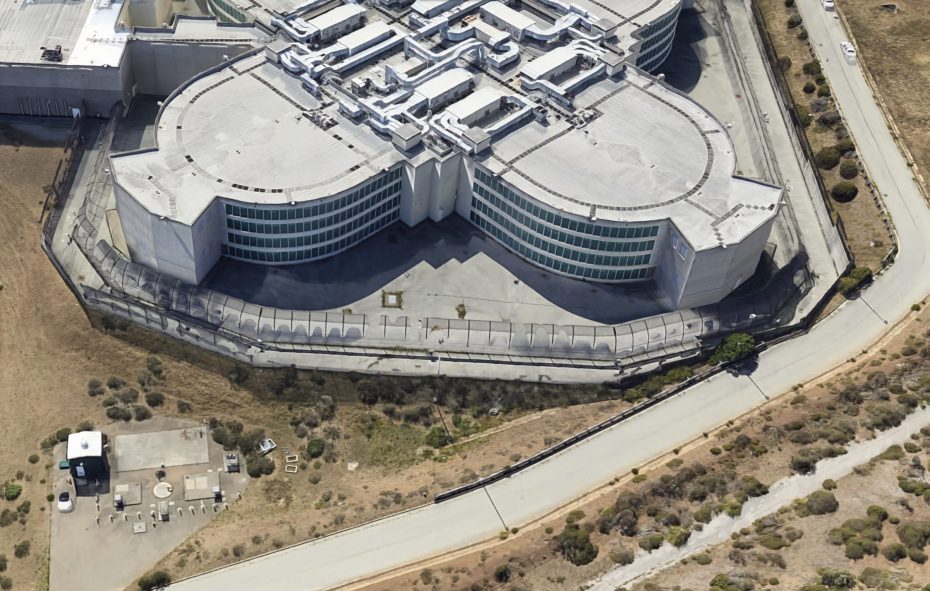 Aerial view of a large, modern, multi-winged building with rounded sections, surrounded by a fence and located near a barren landscape and road.
