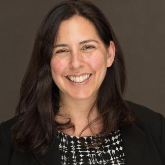 A person with long dark hair smiles, wearing a black blazer and patterned top, with a plain dark background.