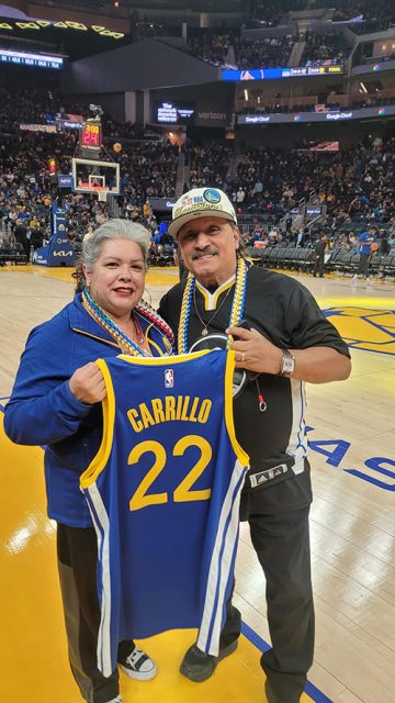 Two people stand on a basketball court holding a jersey with "Carrillo 22" on it. The arena is filled with spectators.