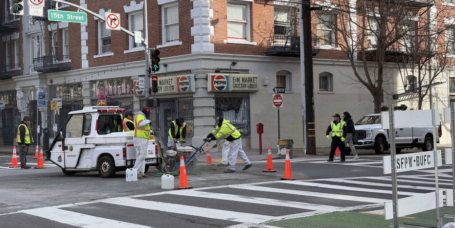 Workers in reflective vests operate machinery at a street intersection with orange cones and traffic control signs. A utility vehicle and truck are parked nearby.