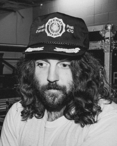 Person with long hair and a beard wearing a cap with a logo and "Post 470" text, looking off-camera. Black and white image.