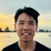 A person smiles warmly outdoors near the water, with the city skyline and a stunning Sutro sunset painting the horizon.