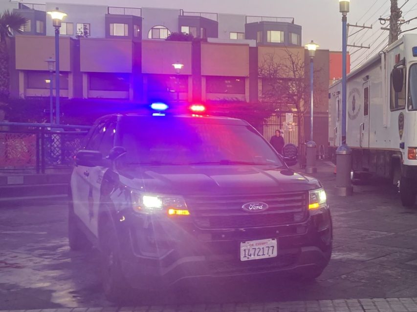 Police SUV with red and blue lights on, parked on a city street at dusk. Background shows buildings and streetlights.