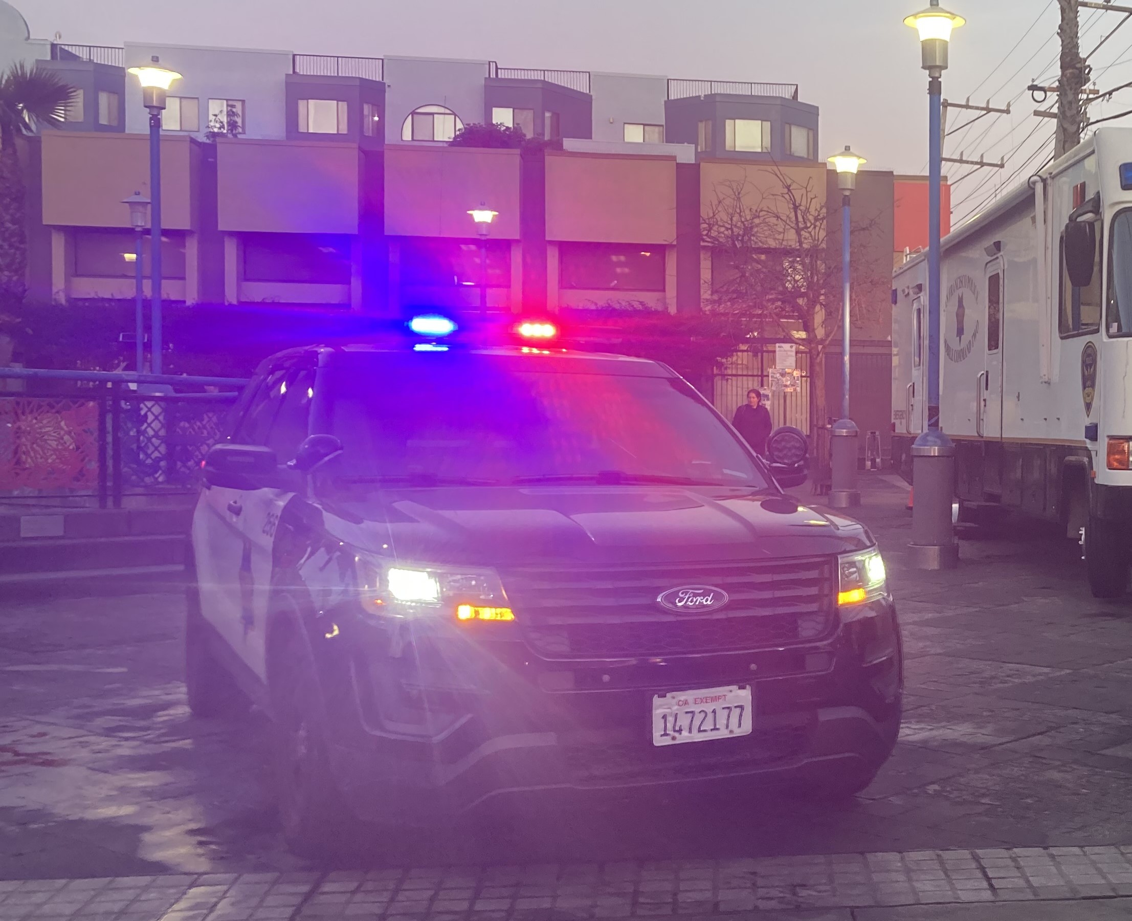 A police SUV with flashing red and blue lights is parked on a street in front of a building. A police van is nearby, and several streetlights are visible.