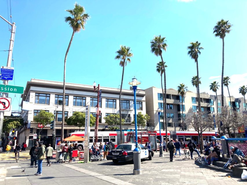 People walking and sitting in a bustling urban plaza with palm trees, buildings, and a police car parked nearby.