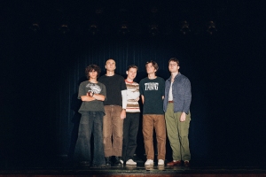 Five people stand together on a dark stage, dressed casually.