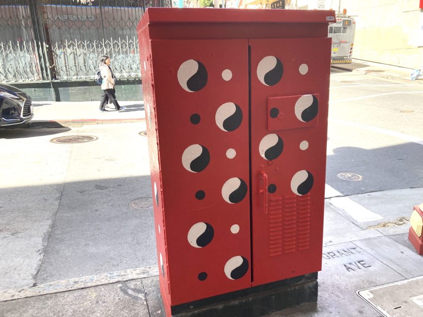A red utility box on a sidewalk, painted with black and white yin-yang symbols. A woman is walking in the background, and a bus is visible on the right side.
