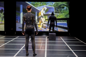 Two people in motion capture suits face a large screen displaying a digital scene with a yellow robot.