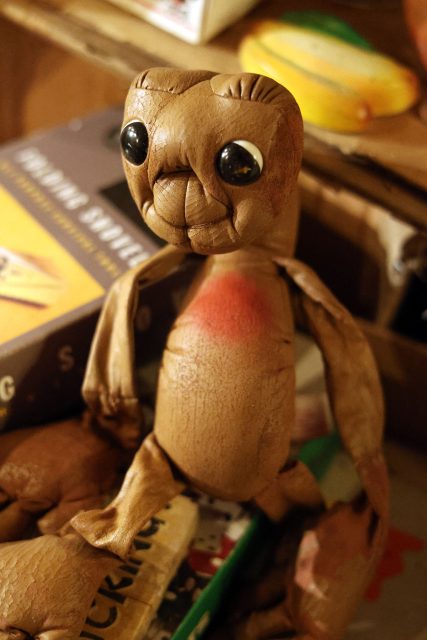 A handmade wooden creature with large black eyes and brown textured body sits on a cluttered surface, surrounded by books and papers.