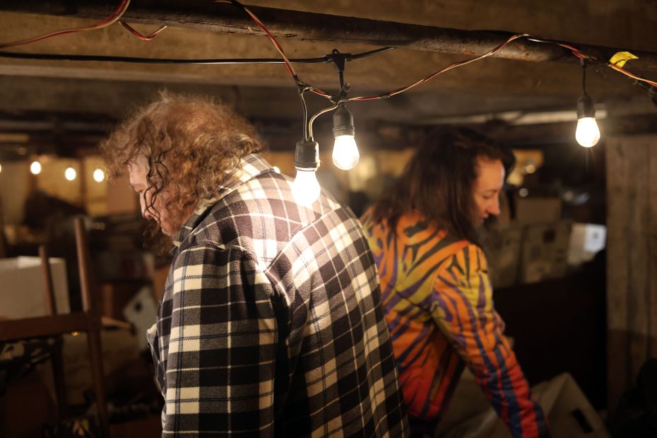 Two people in a dimly lit room with exposed bulbs. One wears a checkered shirt, the other a colorful patterned jacket. Surrounded by miscellaneous items.