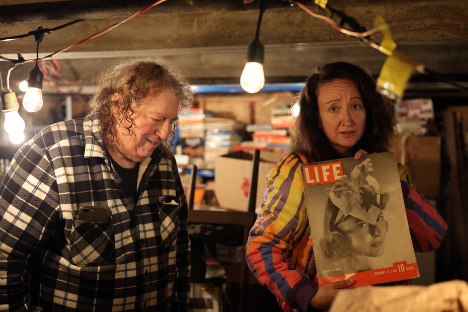 Two people stand in a cluttered basement. The person on the right holds a vintage Life magazine with a woman's face on the cover. Exposed bulbs hang above.