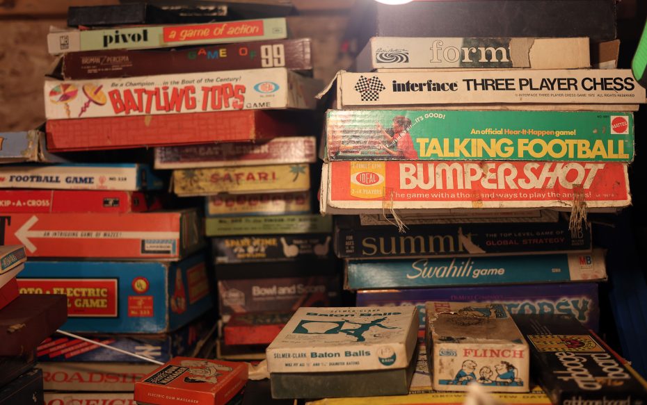 A cluttered pile of vintage board game boxes, including titles like "Talking Football," "Three Player Chess," and "Battling Tops," stacked under a dim light.