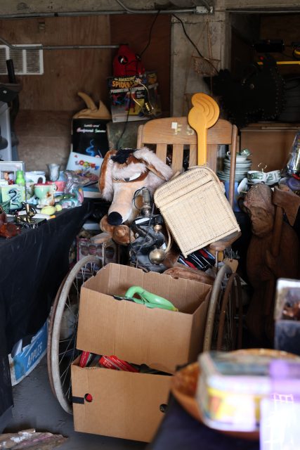 A cluttered room featuring a wheelchair with a large plush toy, a wicker basket, and assorted items. Cardboard boxes filled with miscellaneous objects are in the foreground.