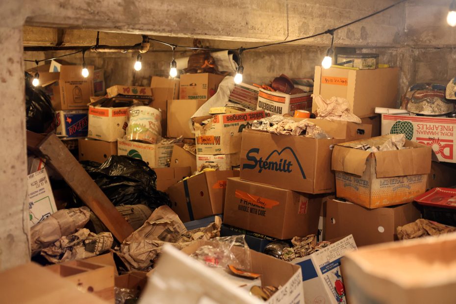 A cluttered basement filled with numerous boxes, newspapers, and bags. Overhead string lights illuminate the space.