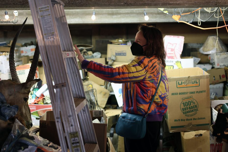 Person in a colorful jacket and mask browsing items in a cluttered basement with boxes, a ladder, and a mounted animal head.