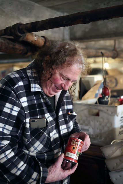 Person in a plaid jacket smiles while holding a can of chili beans. They appear to be in a basement with visible pipes and various items in the background.