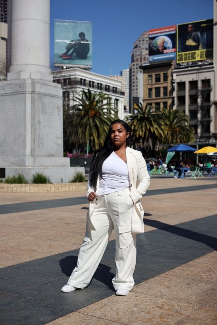 A person stands confidently in a white outfit in a city square, with palm trees and tall buildings in the background under a clear blue sky.