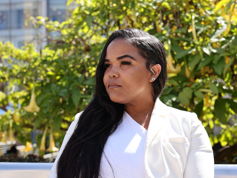 A woman in a white outfit sits outdoors in front of leafy green plants, looking away from the camera.