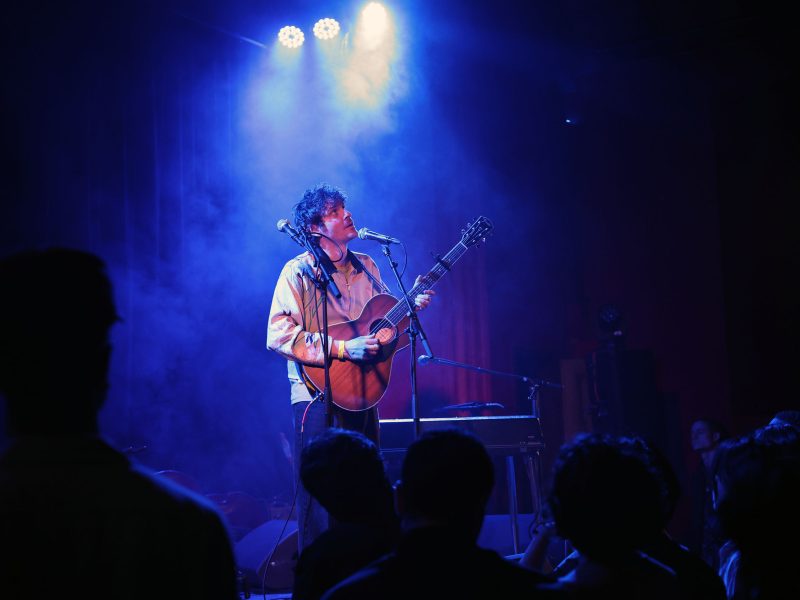Musician plays guitar on stage under blue lights, surrounded by audience in silhouetted foreground.