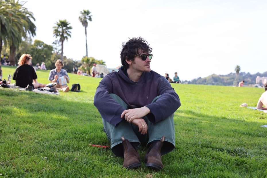 Man in sunglasses and dark hoodie sits on grass in a park, with people and palm trees in the background.
