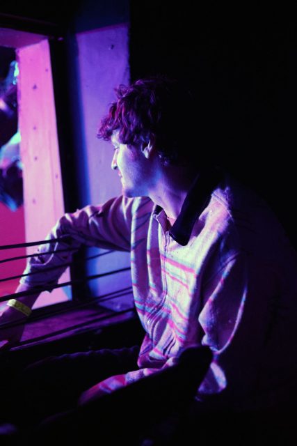 A person in a striped shirt sits in a dimly lit room, illuminated by purple and blue lights, looking towards a window.