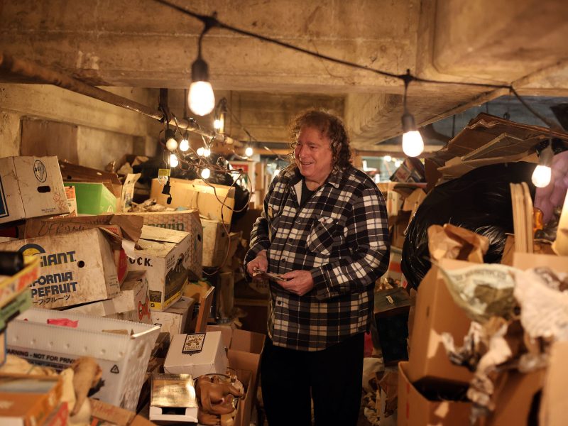 Person standing in a cluttered basement filled with boxes and bags, wearing a plaid shirt. Hanging string lights illuminate the space.
