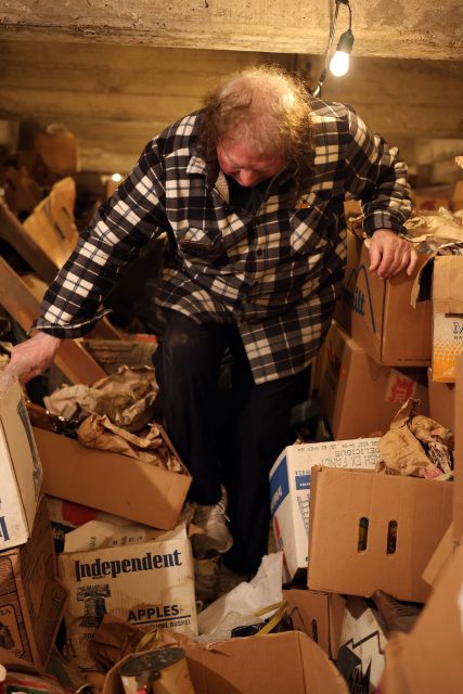 Person in a plaid shirt stands among cluttered boxes and paper in a dimly lit space with a single light bulb above.