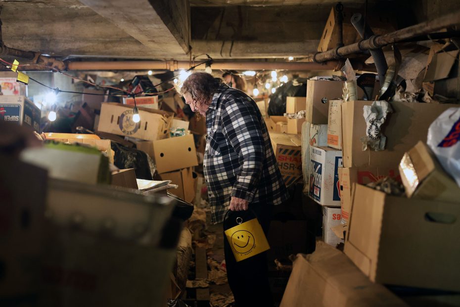 Person in a plaid shirt holds a yellow bucket with a smiley face, standing in a cluttered basement filled with boxes and various items. Dim lighting and exposed pipes are visible.