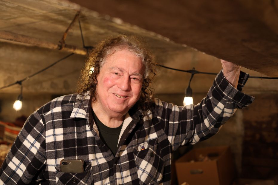 Person with curly hair in plaid shirt stands in a dimly lit room, resting a hand on a wooden beam. Hanging lights illuminate the space.