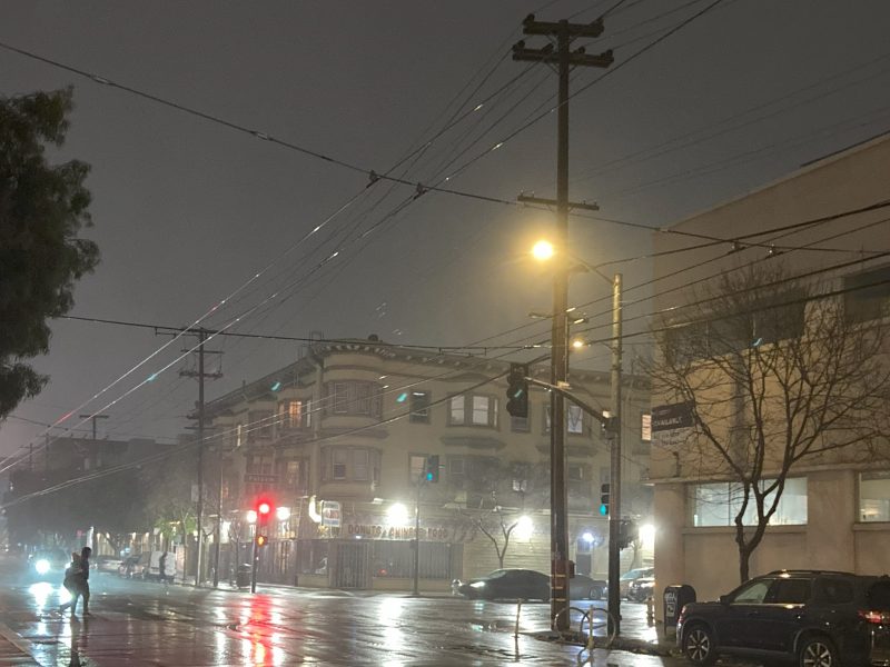 Nighttime city street scene in the rain, with wet pavement reflecting streetlights and traffic signals. A person holding an umbrella walks on the sidewalk near parked cars.