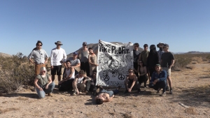 A group of people stand in a desert landscape holding a banner that reads "Bike Plane." One person lies in front. The banner features a small plane illustration and peace symbols.