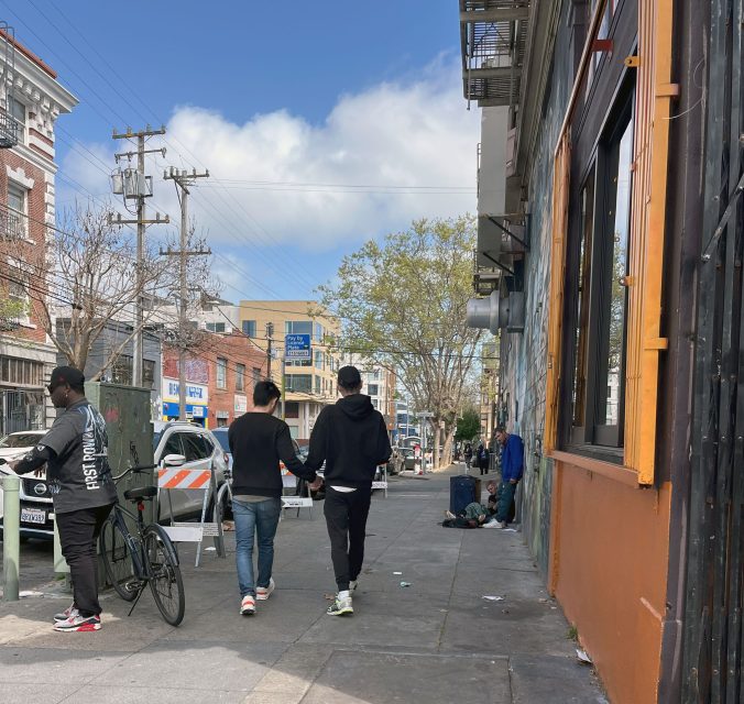 Two people walk down a city sidewalk holding hands. A person on a bicycle is nearby, and some individuals are seated further down the street. Buildings and a traffic barrier are visible.