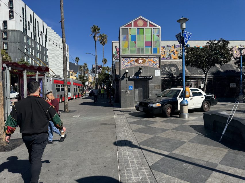 Street scene with pedestrians, a parked police car, a red bus, and a colorful building facade. Palm trees and graffiti are visible under a clear blue sky.