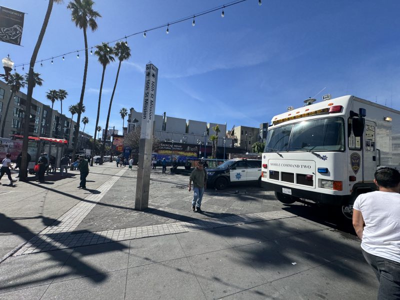 Street scene with palm trees, a police truck, and officers near a busy urban area. People walk nearby under a clear blue sky.
