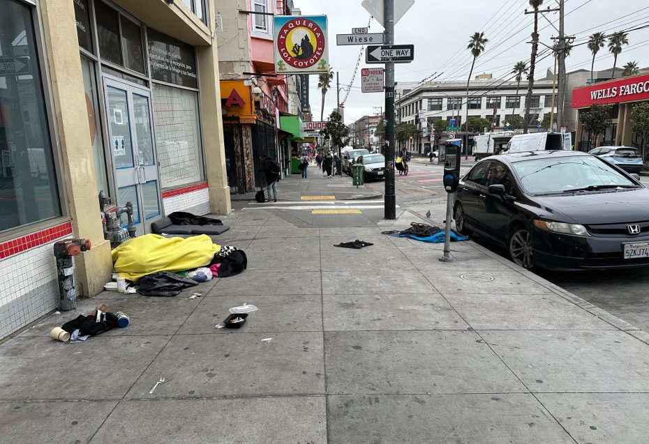 Urban street scene with scattered belongings and a yellow sleeping bag on the sidewalk. Cars are parked on the street, and pedestrians are in the distance near shopfronts.