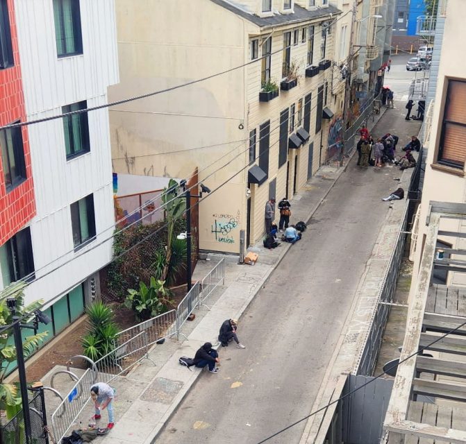 A group of people are gathered along a narrow urban alleyway near buildings, with some sitting, standing, or lying on the ground. Rails and graffiti are visible.