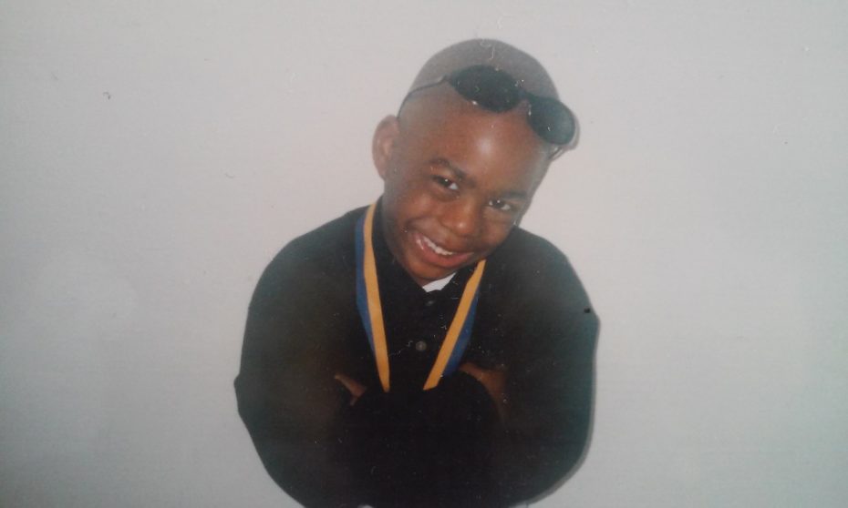 Young boy smiling, wearing a black shirt, blue and yellow medal, and sunglasses on his head, standing against a plain background.