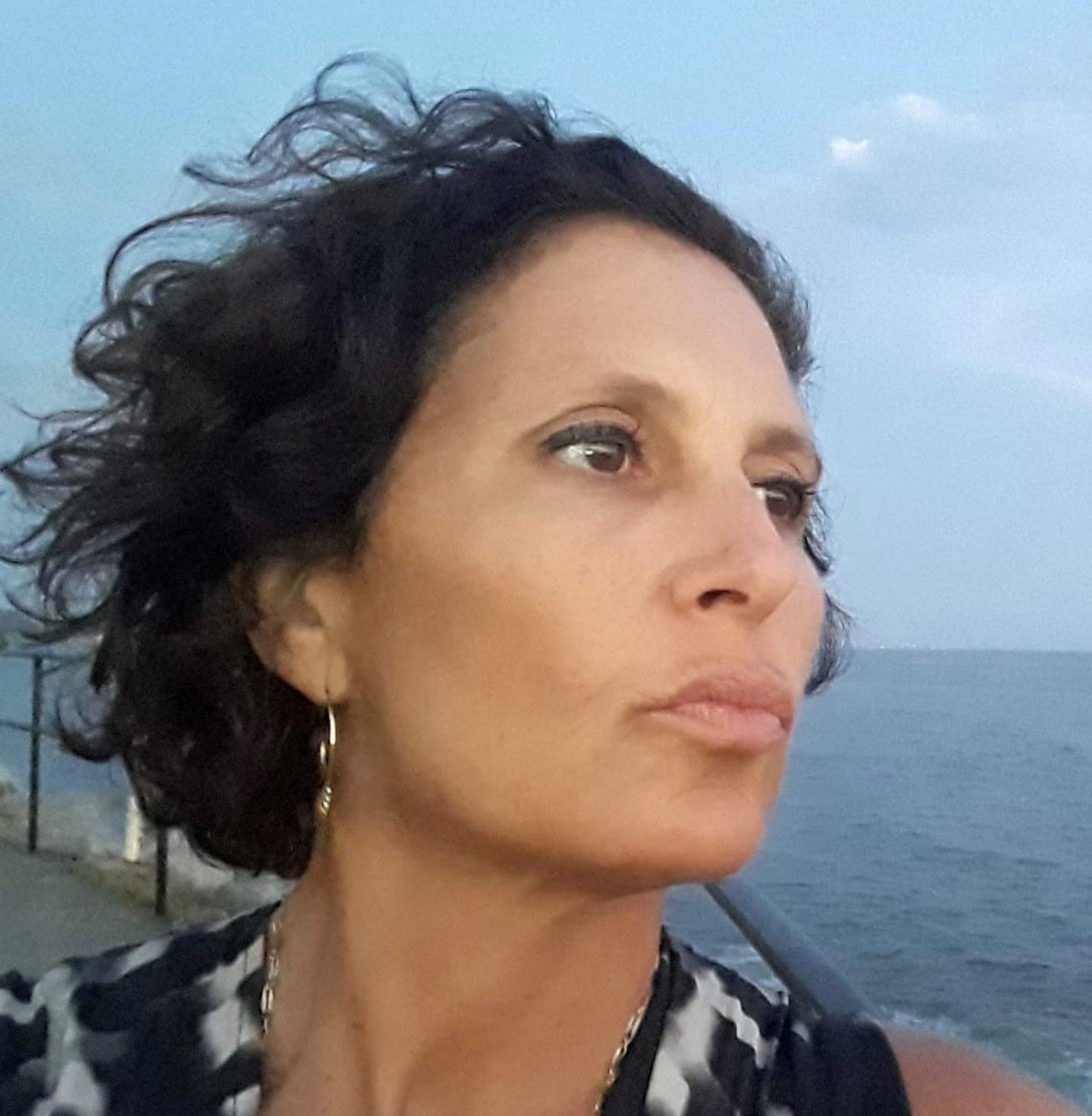 Woman with curly hair looking into the distance by the sea, wearing a patterned top and hoop earrings.