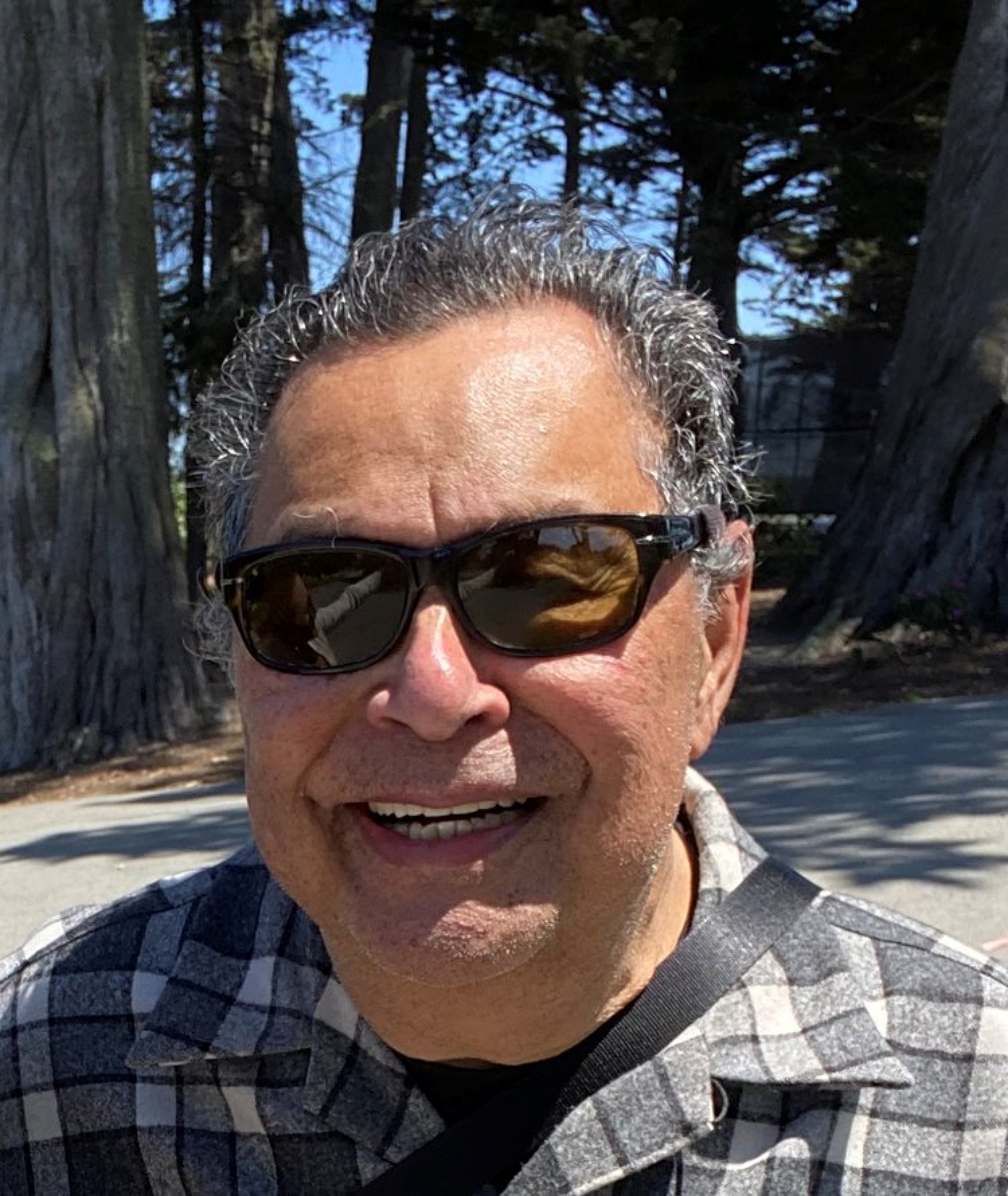 Mission filmmaker Ray Balberan with sunglasses smiles outdoors, wearing a checkered shirt. Trees and a clear sky are in the background.
