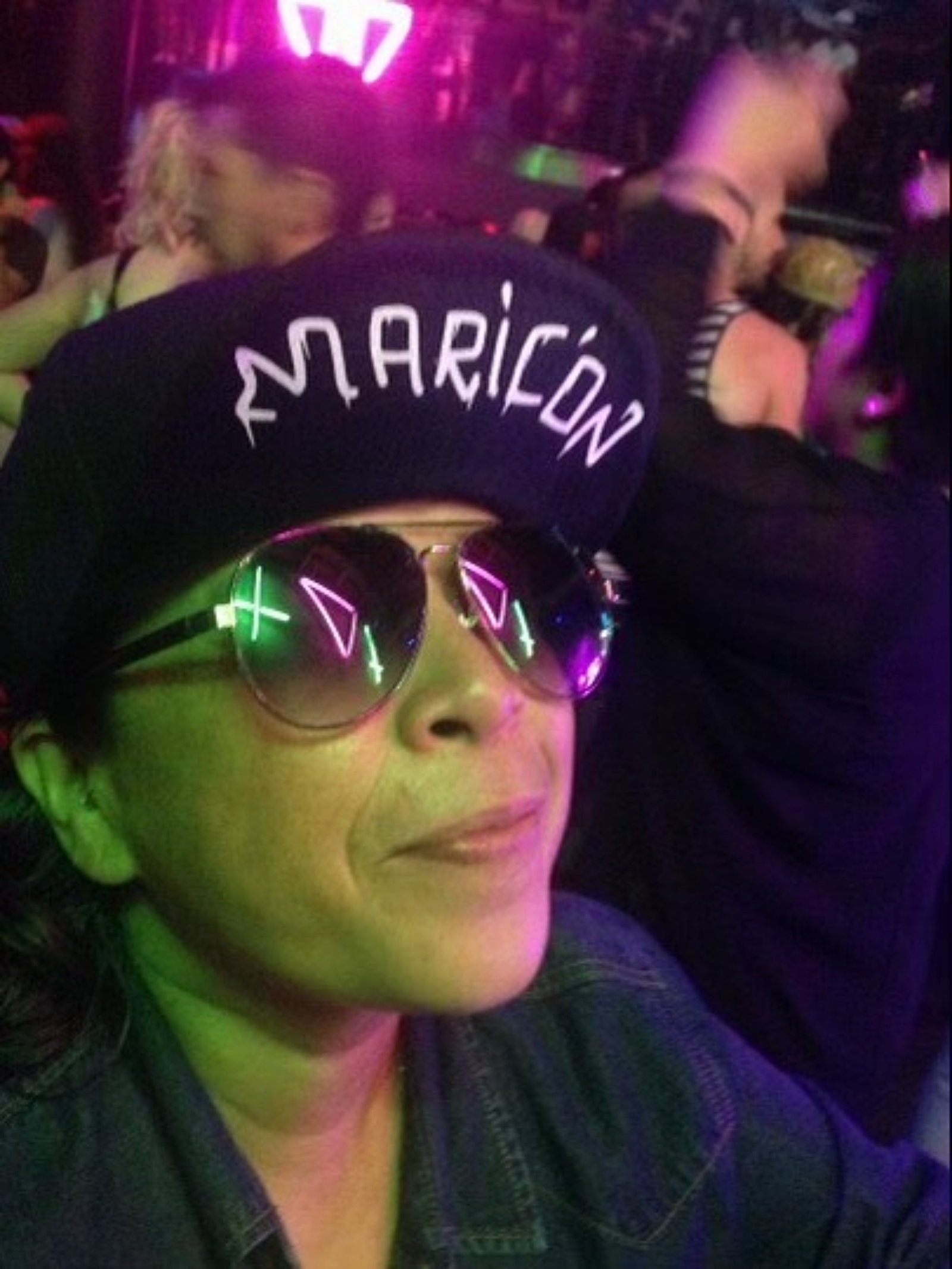 In the vibrant glow of colorful lights, a person wearing sunglasses and a cap emblazoned with "MARICON" stands amidst the bustling crowd, as if on a mission to merge style with individuality.