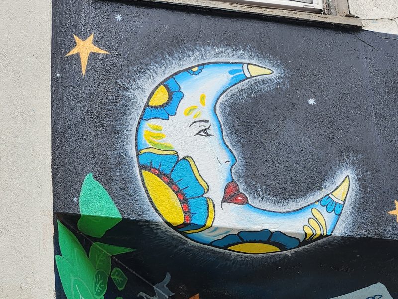 Colorful mural of a crescent moon with a face and floral patterns against a starry night background on a wall.